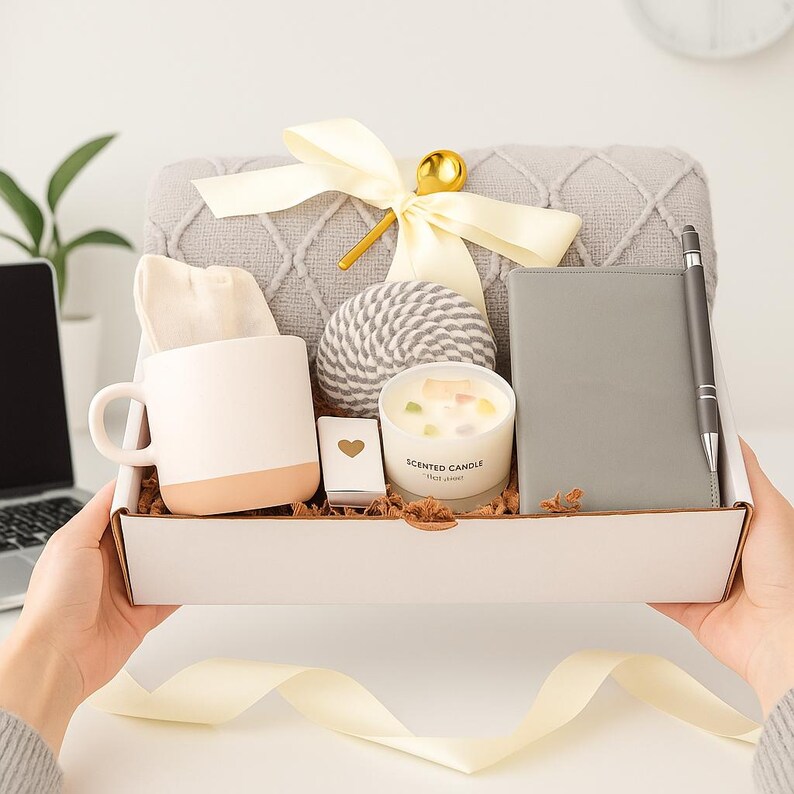 Puede incluir: Una caja de regalo llena de una manta gris, una taza blanca y beige, una vela, un cuaderno, un bol&iacute;grafo, una bola decorativa y una cajita con un coraz&oacute;n. La caja est&aacute; atada con una cinta color crema.