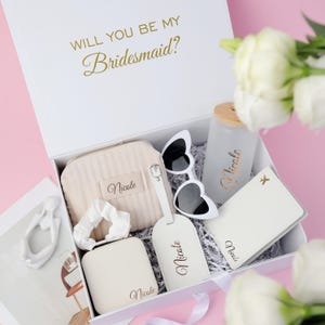 May include: A white gift box with gold text reading "WILL YOU BE MY Bridesmaid?" contains a variety of items. These include a beige cosmetic bag, heart-shaped sunglasses, a water bottle, a luggage tag, and a passport holder. All items are personalized with the name "Nicole".