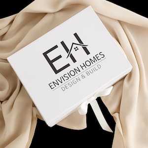 May include: A rectangular white gift box featuring a black logo with the text ENVISION HOMES DESIGN & BUILD and a graphic of a house roof. The box is closed with a white ribbon tied in a bow, resting on a draped beige fabric background.