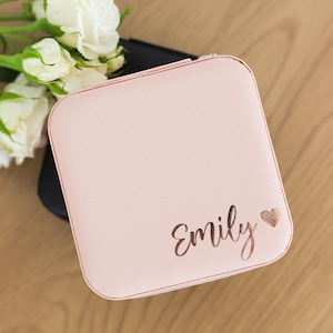 May include: A square, light pink jewelry box with a textured finish. The lid features the name Emily written in a metallic rose gold script font, accompanied by a small, matching rose gold heart icon positioned to the right of the name.