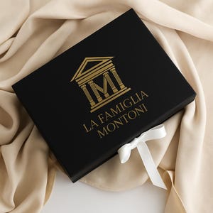 May include: A rectangular black gift box featuring a gold-colored logo of a classical temple structure above the text LA FAMIGLIA MONTONI. The box is accented with a white satin ribbon tied in a bow on the lower right corner of the lid.