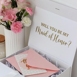 May include: A white gift box with the gold text Will You Be My Maid of Honor? printed on the inside lid. The box contains white paper shred filler, a light pink envelope, and a white card with black polka dots and gold script text.