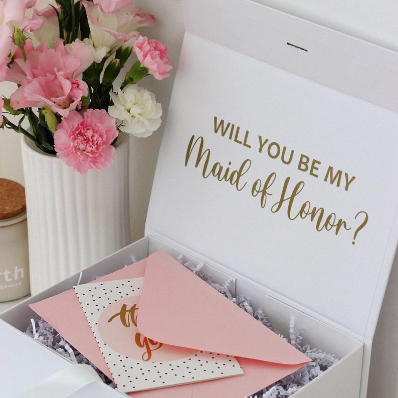 May include: A white gift box with the gold text Will You Be My Maid of Honor? printed on the inside lid. The box contains white paper shred filler, a light pink envelope, and a white card with black polka dots and gold script text.