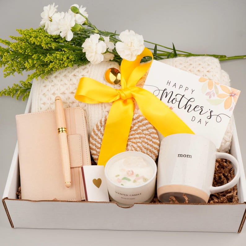 Puede incluir: Una caja de regalo para el D&iacute;a de la Madre, que contiene una taza blanca con la palabra "mam&aacute;" impresa, una vela, un cuaderno, un bol&iacute;grafo, una tarjeta y una cinta amarilla. La caja tambi&eacute;n incluye una manta de punto blanca, un posavasos y flores frescas.