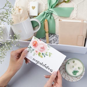 Puede incluir: Una caja de regalo con una taza blanca, un cuaderno peque&ntilde;o, un bol&iacute;grafo, una vela y una tarjeta que dice "felicidades". La tarjeta tiene rosas de acuarela. La caja est&aacute; atada con una cinta verde.