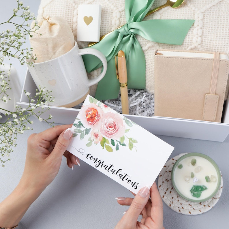 Puede incluir: Una caja de regalo con una taza blanca, un cuaderno peque&ntilde;o, un bol&iacute;grafo, una vela y una tarjeta que dice "felicidades". La tarjeta tiene rosas de acuarela. La caja est&aacute; atada con una cinta verde.