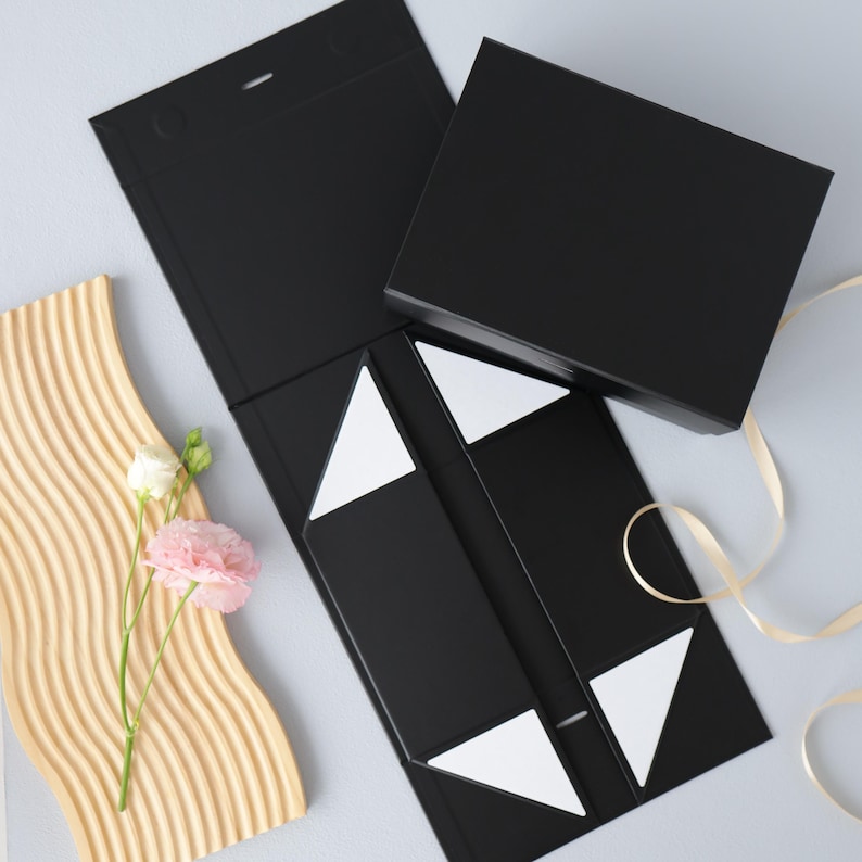 May include: A black rectangular gift box with a magnetic closure lid, shown in both open and closed positions. The open box features white triangular adhesive tabs on the interior flaps. A light beige ribbon and a pink flower are nearby.