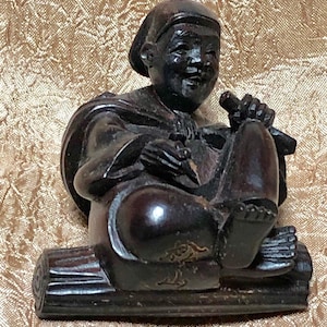May include: A dark brown wooden figurine of a person sitting on a log. The person is wearing a hat and a robe. The figurine is carved with intricate details.