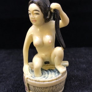 以下が含まれることがあります： 長い黒髪の女性が木製の浴槽に座っている、小さな象牙の置物。女性は裸で、顔には穏やかな表情が浮かんでいる。浴槽は波模様で飾られている。