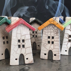 May include: A collection of ceramic house-shaped incense burners with white crackle-glazed walls and dark gray arched doors. Each house features a distinct colored roof in red, orange, green, or blue, with wisps of smoke rising from the chimneys.