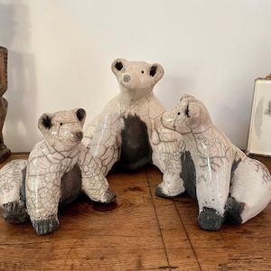 May include: Three ceramic bear ornaments. The bears are white with black detailing on their paws and backs. The ceramic has a crackle glaze. The ornaments are on a wooden surface.