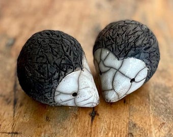 May include: Two ceramic hedgehog figurines. The hedgehogs have a textured, dark gray back and a white face with a cracked pattern. The figurines are set on a wooden surface.