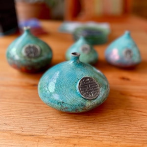 May include: A collection of turquoise ceramic vases with a crackle glaze. Each vase has a round shape with a small opening at the top and a circular emblem on the side. The vases are arranged on a wooden surface.