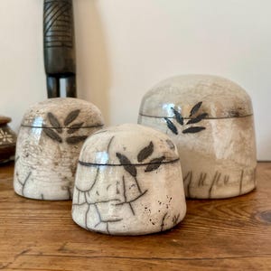 Peut inclure: Trois pots en céramique en forme de dôme avec couvercle, dotés d'une finition émaillée craquelée couleur crème. Chaque pot présente un motif de feuilles peint à la main en brun foncé et une ligne horizontale. La surface montre un réseau de craquelures noires.