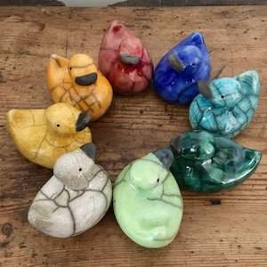 May include: A collection of seven small ceramic duck figurines in various colors, including yellow, red, blue, green, and white. Each duck has a cracked glaze finish, giving them a rustic and unique look.