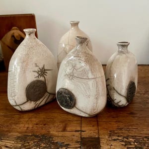 May include: Four handcrafted ceramic vases with a speckled, off-white glaze. Each vase has a unique shape and features dark, textured accents and a circular, dark-colored embellishment. The vases are arranged on a wooden surface.