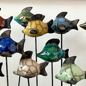 Könnte beinhalten: Eine Sammlung von Keramikfischskulpturen in verschiedenen Farben, darunter Rot, Blau, Gelb und Grün. Jeder Fisch hat eine dunkle Flosse und ist auf einem schwarzen Metallstab montiert. Die Fischskulpturen sind in einer dekorativen Anordnung angeordnet.