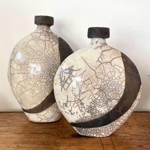 Puede incluir: Dos jarrones de cerámica con un esmalte blanco craquelado y un diseño de rayas negras. Los jarrones son redondos y tienen una abertura ancha en la parte superior.