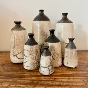 Könnte beinhalten: Ein Set aus sieben Keramikvasen in verschiedenen Größen, alle mit einer weißen Craquelé-Glasur und schwarzen Rändern. Die Vasen sind auf einer Holzoberfläche angeordnet.
