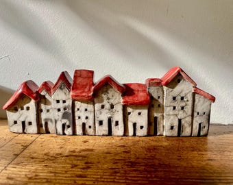 May include: A ceramic sculpture featuring a row of seven small houses with bright red roofs and off-white walls. Each house has a unique shape, small square windows, and dark crackle patterns across the surface, creating a continuous village scene.