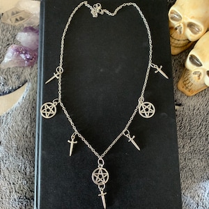 May include: A silver chain necklace with five charms: two pentagram charms, three sword charms. The necklace is on a black background.