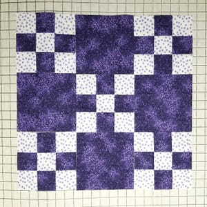 May include: A square quilt block featuring a nine-patch pattern composed of alternating fabric squares. The design combines solid dark purple floral print fabric with white fabric squares featuring a small purple star-like pattern throughout.