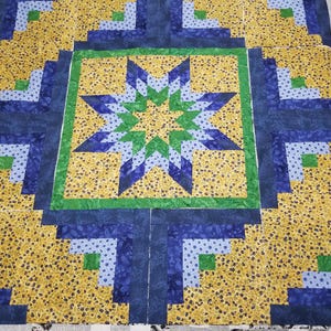 May include: A quilt featuring a central star pattern in shades of blue, green, and yellow. The star is surrounded by a green border and log cabin blocks in yellow, light blue, and dark blue fabric with various floral and leaf patterns.