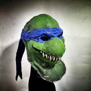 Puede incluir: Una máscara de las Tortugas Ninja de color verde y azul con textura de arpillera y una máscara para los ojos azul. La máscara tiene una boca cosida con dientes afilados.