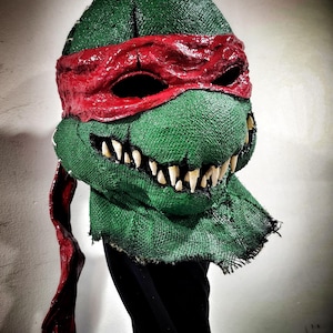 Puede incluir: Una máscara verde y roja de las Tortugas Ninja con una textura de tela deshilachada y una sonrisa amenazante. La máscara tiene una bandana roja que cubre los ojos y una fila de dientes afilados.