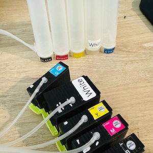 May include: Six ink cartridges for a printer, including black, cyan, magenta, yellow, and white. The cartridges are labeled with their colors and have clear tubes attached to them.