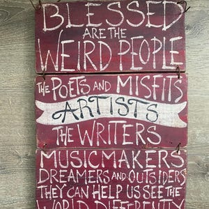 May include: A rustic, wooden sign with a burgundy background and white lettering. The sign is composed of three stacked planks, each bearing inspirational text about creative individuals, including poets, artists, and dreamers. A wire is attached for hanging.