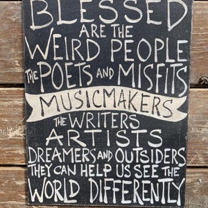 May include: Black wooden sign with white text that reads "Blessed are the weird people the poets and misfits musicmakers the writers artists dreamers and outsiders they can help us see the world differently."