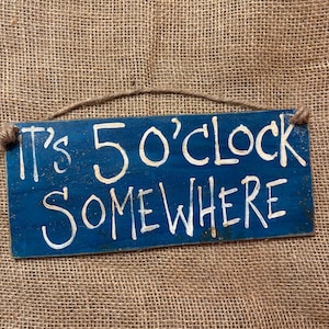 May include: A rectangular blue wooden sign with the text "It's 5 O'Clock Somewhere" in white lettering. The sign is suspended by a natural twine rope. The background is a textured burlap.