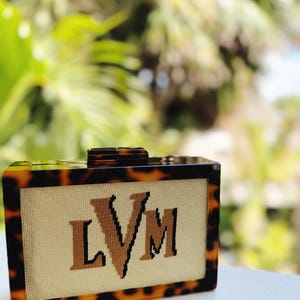 May include: A rectangular box clutch with a brown and black tortoiseshell pattern frame. The center panel features a cream-colored fabric with the letters LVM embroidered in brown and black thread. A matching tortoiseshell clasp sits on top.