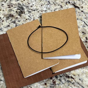 May include: A set of kraft paper notebooks with white paper pages, bound with a black elastic band. The notebooks are stacked on top of a brown leather-like cover, creating a layered effect. The background is a speckled, light-colored surface.