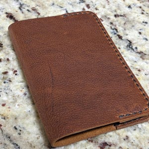 May include: A brown leather wallet with visible stitching around the edges. The wallet has a textured surface and appears to be made of high-quality leather. The wallet is rectangular and has a folded design.