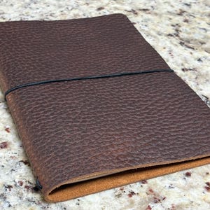 May include: A closed, brown leather journal with a textured surface and a black elastic band closure. The edges of the journal are a lighter brown color. The journal is lying flat on a speckled surface.