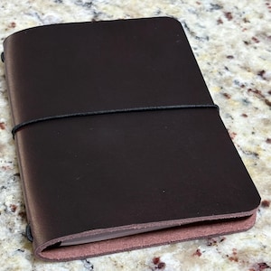 May include: A dark brown leather notebook cover with rounded corners, secured by a thin black elastic band wrapped horizontally around the center. The cover features a smooth, matte finish and shows the raw, textured edge of the leather.