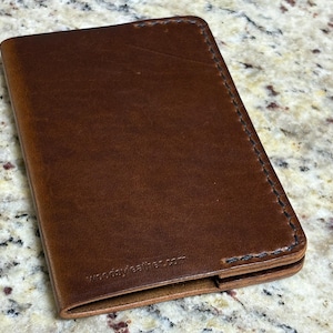 May include: A brown leather wallet with visible stitching along the edges. The wallet has a smooth surface and a slightly worn appearance. The website address "woodyleather.com" is subtly embossed on the bottom corner. This wallet is designed for carrying cards and cash.
