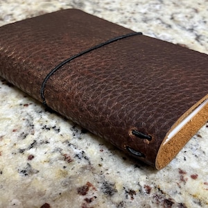 May include: A brown leather journal with a black elastic closure. The journal has a textured surface and is closed.