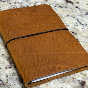 May include: A closed, rectangular, brown leather journal with a black elastic band closure. The journal has a textured surface and visible stitching along the edges. The pages are white. The journal is lying on a speckled surface.