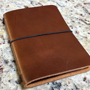 May include: A brown leather journal with a black elastic band closure. The journal is rectangular and appears to be closed. The leather has a natural grain and texture. The journal is likely for writing or sketching.