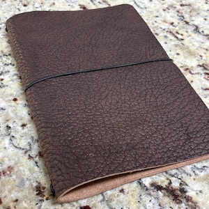 May include: A brown leather journal with a textured surface and a black elastic band closure. The journal is rectangular and appears to be closed. The edges are rounded. The journal is on a speckled surface.