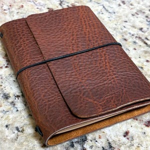 May include: A brown leather journal cover with a textured, pebbled surface. The cover features a wrap-around design secured by a thin black elastic band that stretches horizontally across the front, holding the folded leather flap in place.