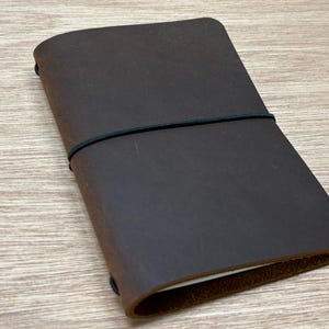 May include: A dark brown leather journal cover with rounded corners, featuring a smooth matte texture and a single black elastic band wrapped horizontally around the center to keep the notebook securely closed.