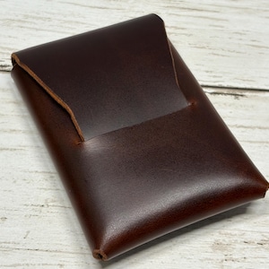 May include: A dark brown leather card holder with a smooth finish and a single flap closure. The rectangular accessory features a simple folded construction with visible edges and a deep, rich color tone throughout the entire surface area.