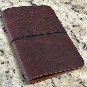 May include: A closed, rectangular, dark brown leather journal with a black elastic band closure. The leather has a textured appearance. The journal is lying on a speckled, light-colored surface.