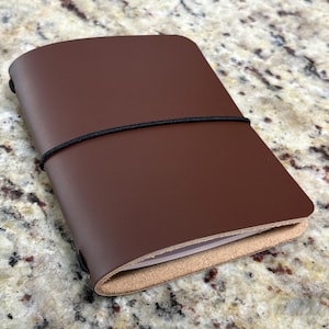 May include: A brown leather notebook cover with a smooth, matte finish and rounded corners. The cover is secured by a thin black elastic band that wraps horizontally around the center, holding the folded leather structure closed over white paper pages.