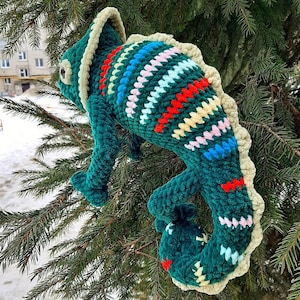 May include: A plush crochet chameleon toy in a deep green hue, featuring a textured cream-colored crest along its back and head. The body is decorated with horizontal stripes in vibrant red, light blue, yellow, and soft pink yarn accents.