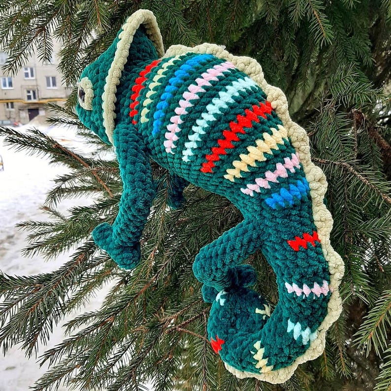 May include: A plush crochet chameleon toy in a deep green hue, featuring a textured cream-colored crest along its back and head. The body is decorated with horizontal stripes in vibrant red, light blue, yellow, and soft pink yarn accents.